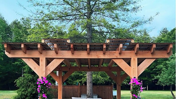 Une pergola en bois, avec un banc et deux chaises en dessous, entourée d’un jardin fleuri et d’un arbre.