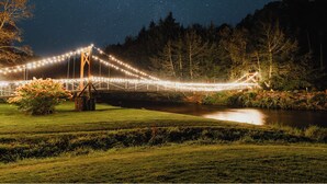 Un pont suspendu illuminé par des guirlandes lumineuses la nuit, entouré d’une forêt et traversé par une rivière.