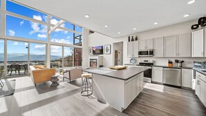 Une cuisine moderne dotée d’un grand îlot central, de meubles blancs et offrant une vue sur les montagnes grâce à de grandes fenêtres.