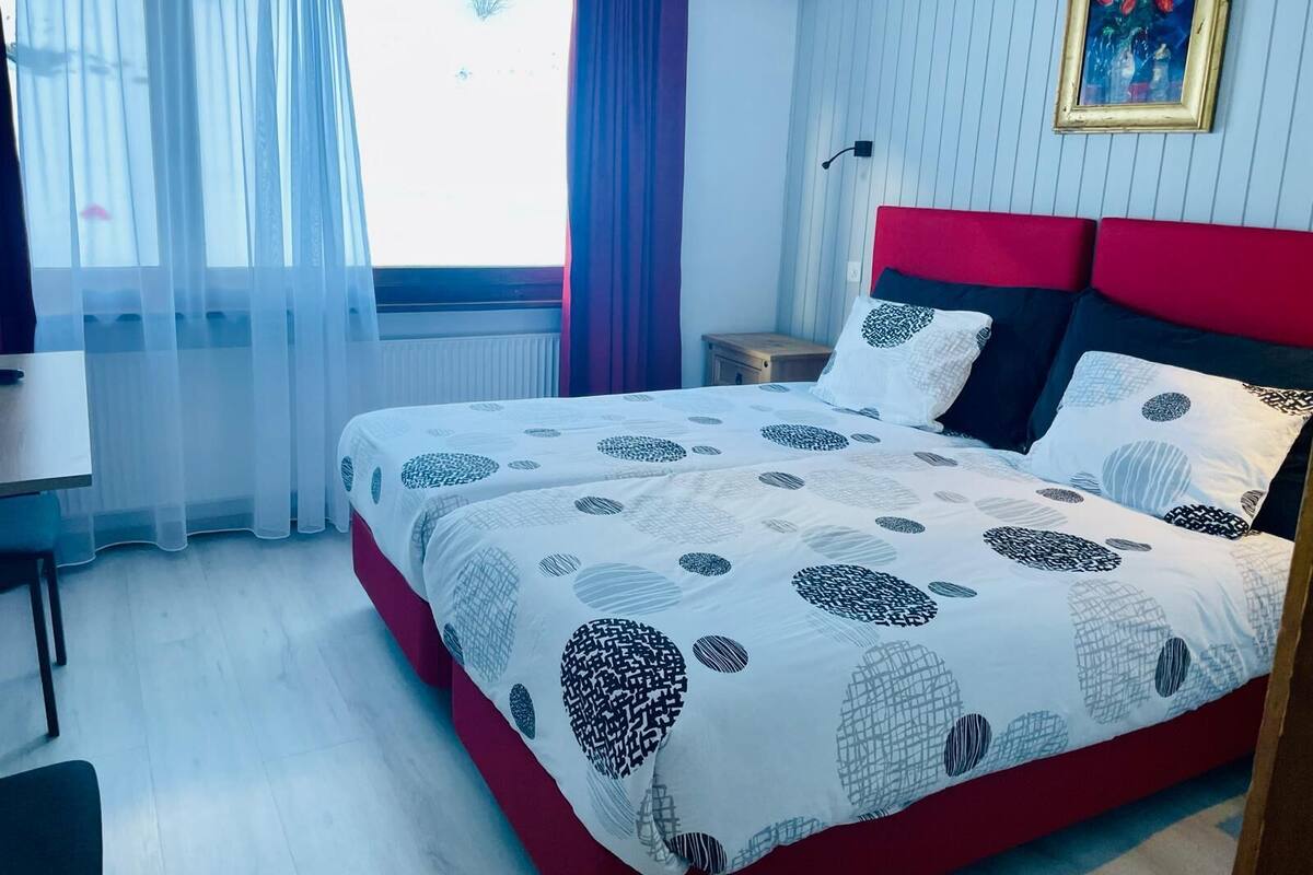 Une chambre à coucher avec un lit, une chaise, une table de chevet, une fenêtre avec des rideaux et un tableau accroché au mur.