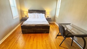 Une chambre avec un plancher en bois, un lit recouvert de draps blancs, deux tables de chevet avec des lampes, un ventilateur de plafond et un bureau avec une chaise.