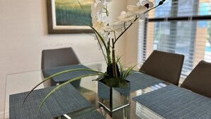 A modern dining area with a glass table and grey upholstered chairs, a vase with white flowers, and a painting on the wall.
