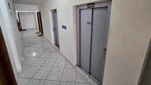 A hallway with tiled flooring, a social elevator, and a door with a sign. - Studio - Av. Presidente. Kennedy  V. Tupi - PG (Vila Tupi Praia Grande)