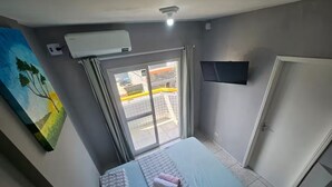 A bedroom with a bed, an air conditioning unit, a TV, and a sliding glass door to the outside. - Studio - Av. Presidente. Kennedy  V. Tupi - PG (Vila Tupi Praia Grande)