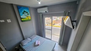 A bedroom with a bed, a painting, an air conditioning unit, a sliding glass door, and a view of buildings outside. - Studio - Av. Presidente. Kennedy  V. Tupi - PG (Vila Tupi Praia Grande)