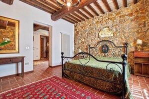 Quarto com parede de pedra, cama com colcha estampada, um banco de madeira e um tapete vermelho estampado.