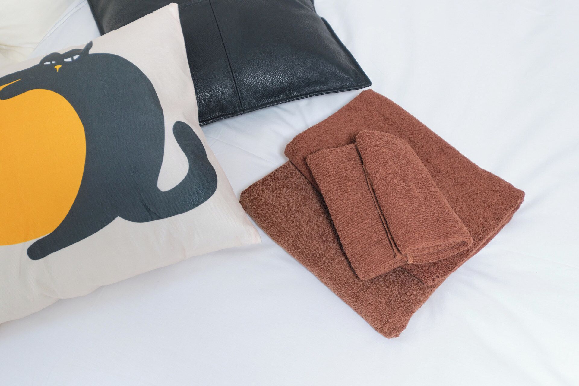 Una cama con un almohadón que lleva un estampado grande de elefante, un almohadón de cuero negro y dos toallas marrones dobladas.