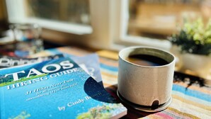 Un libro titulado "TAOS HIKING GUIDE" de Cindy Brown, una taza de cerámica y una ventana con vistas a la vegetación.
