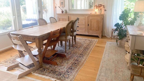 Un comedor con una mesa y sillas de madera, un espejo grande sobre la mesa y vistas al exterior a través de las ventanas.