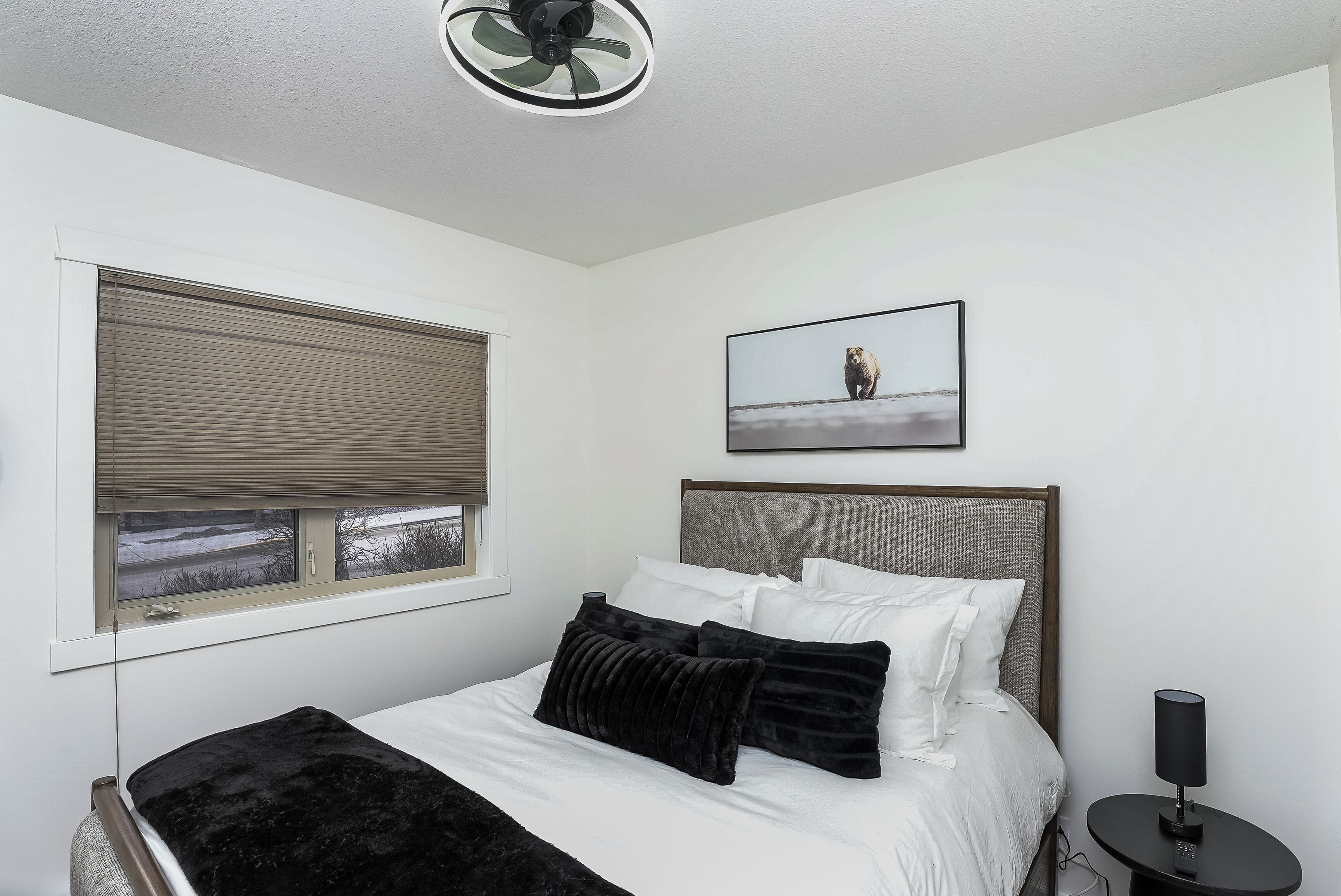 A bedroom with a bed, a window with blinds, a ceiling fan, a bedside table with a lamp, and a framed picture on the wall.