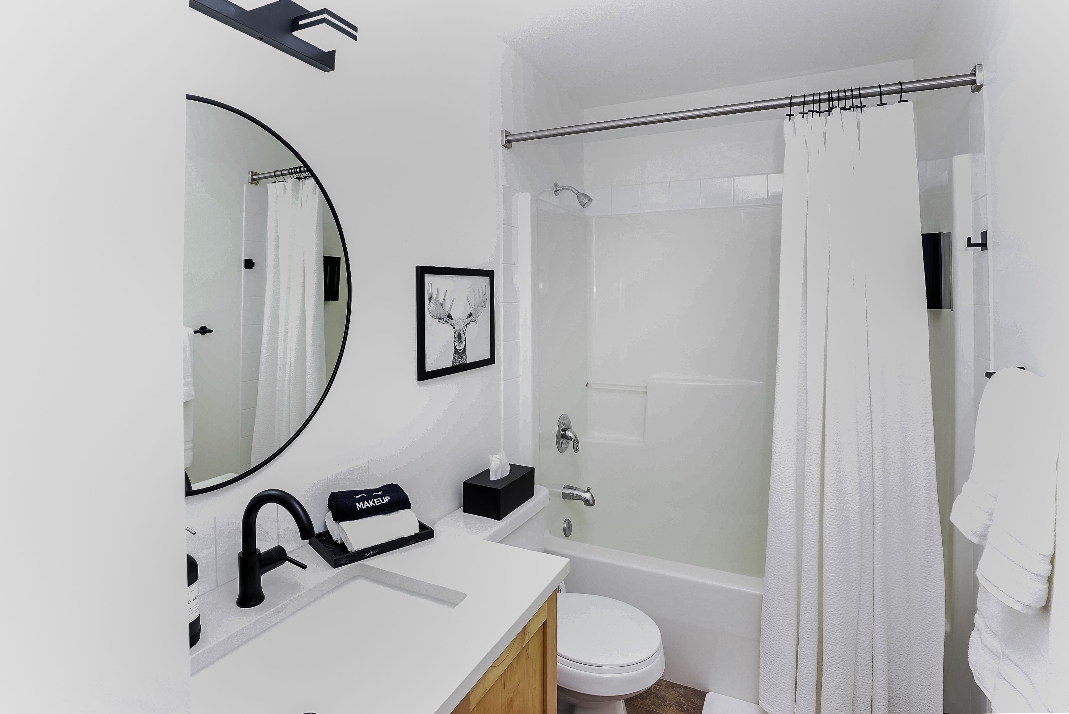 A modern bathroom with a white sink, black faucet, a framed deer picture, a white toilet, and a glass-enclosed shower with a white curtain.