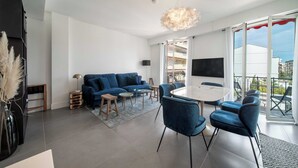 Una sala de estar moderna con un sofá azul, una mesa de comedor con sillas, un televisor grande y un balcón con vistas.