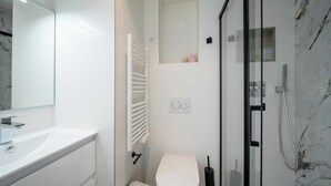 Un baño moderno con un lavabo blanco, un inodoro, cabina de ducha de vidrio y pared de mármol.