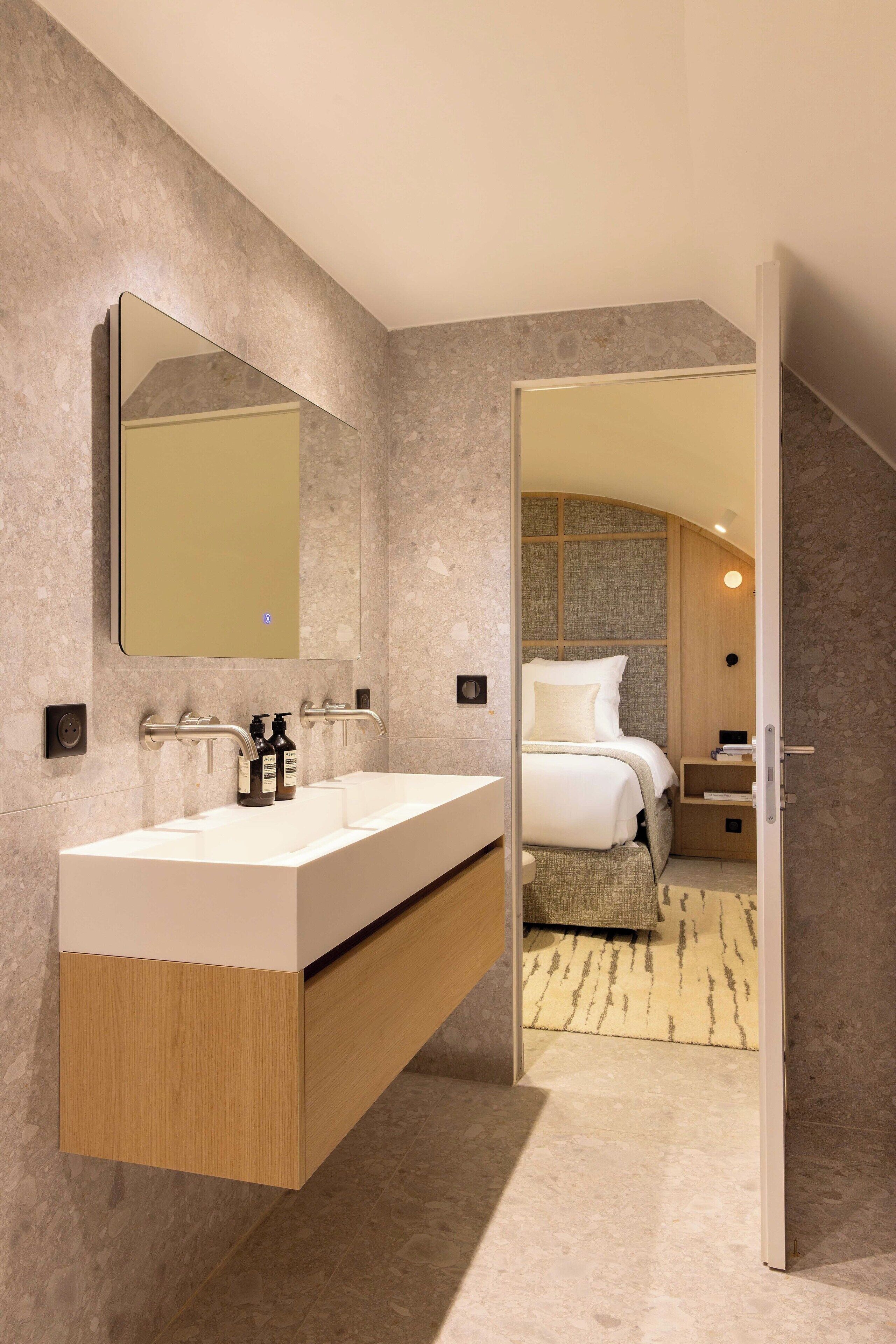 A modern bathroom with a large mirror, a double sink vanity, and a visible bedroom with a bed and nightstand.
