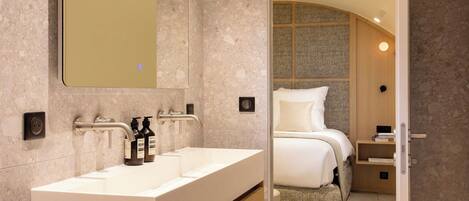 A modern bathroom with a large mirror, a double sink vanity, and a visible bedroom with a bed and nightstand.