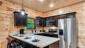 A modern kitchen with wooden walls, stainless steel appliances, granite countertops, and bar stools. - Modern Cabin with Hot Tub, Air Hockey, King Beds, Grill and Outdoor Pool! (Sevierville)