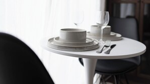 A dining table set with plates, bowls, and cutlery, with a chair in the foreground and a window with curtains in the background.