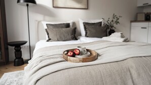 A neatly made bed with a white comforter, a wooden tray with apples and a cup, a floor lamp, and a plant in the corner.
