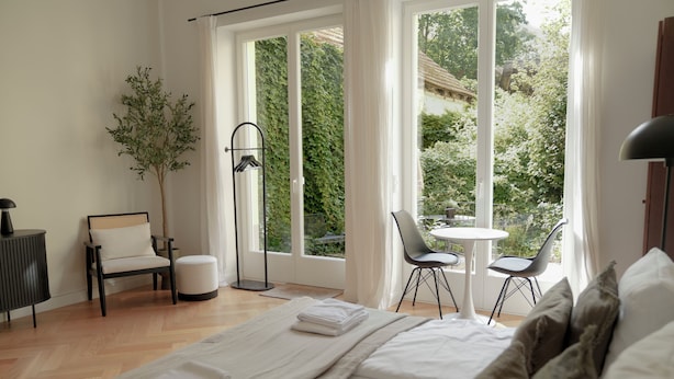A modern bedroom with a large window, a bedside table, a chair, and a small table.