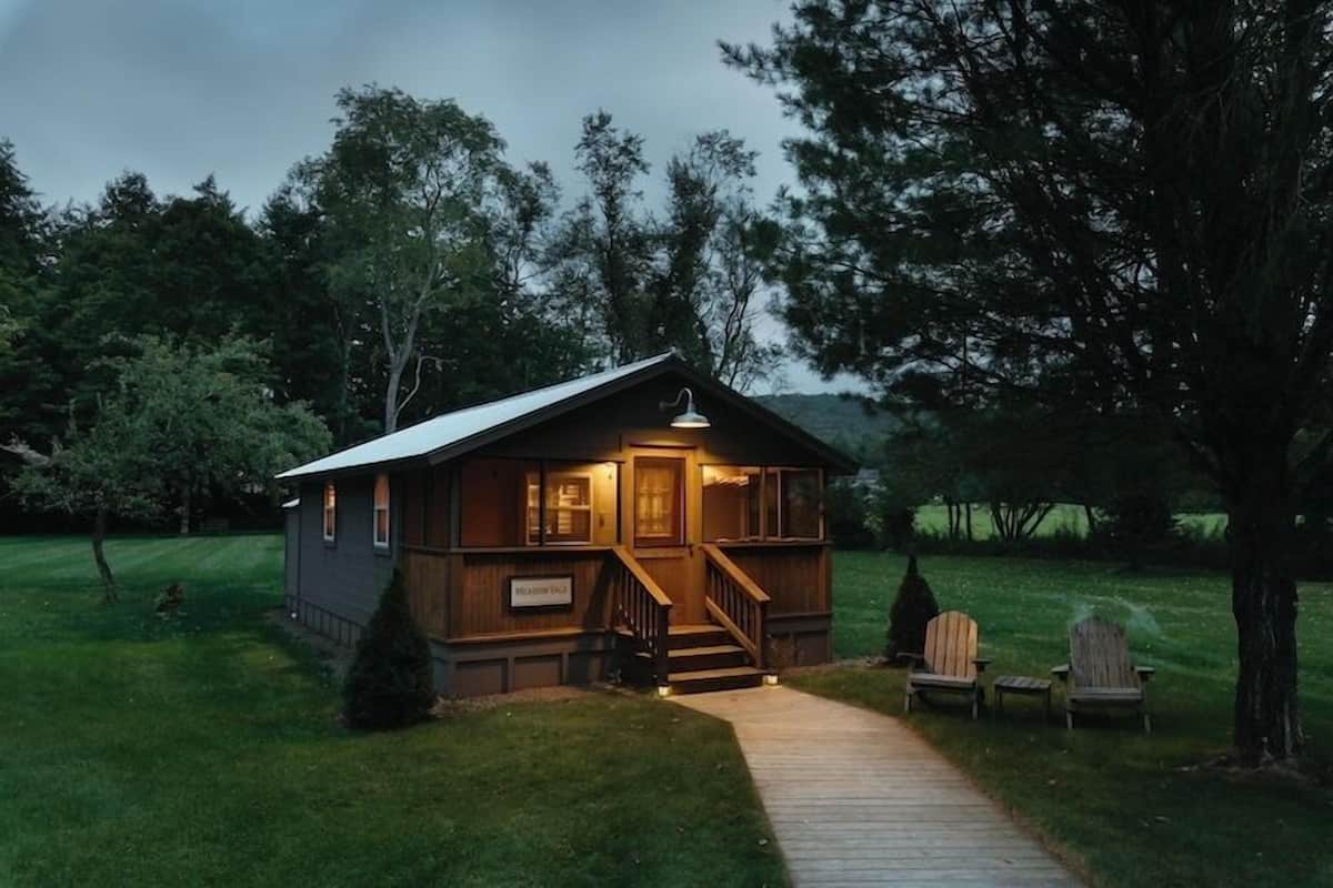 A small cabin with a porch and steps, surrounded by trees and a grassy area.