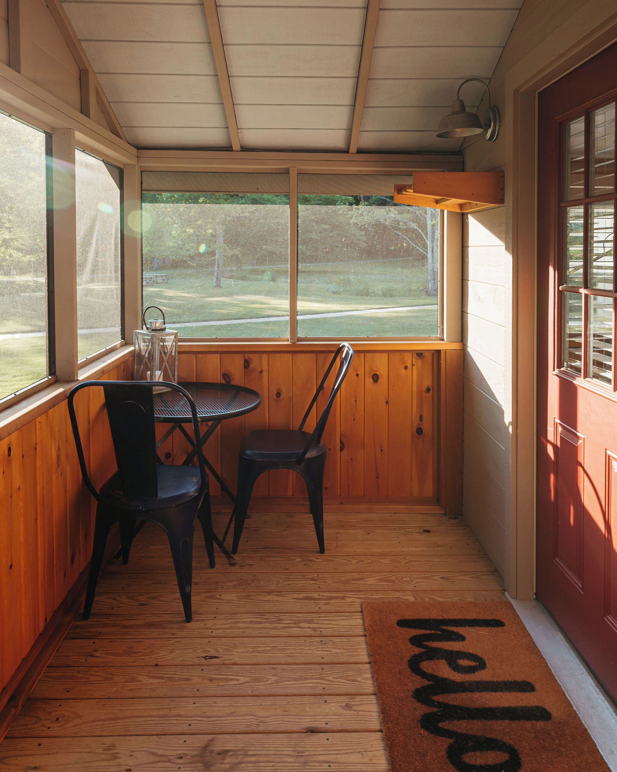 A wooden porch with a table and two chairs, a 'hello' mat, and a view of a grassy area through large windows.