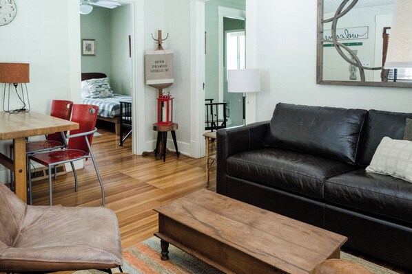 A living room with a leather sofa, a wooden coffee table, and a red chair. There is a bed with a striped bedspread in the background.
