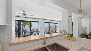 A modern kitchen with a large window overlooking a pool and ocean view, a ceiling fan, and a hanging light fixture.