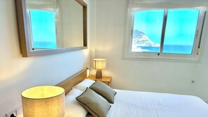 Una cama bien tendida con colcha blanca, dos almohadas y una lámpara de noche sobre una mesita de madera. Un espejo grande cuelga sobre la cama, y se ve una ventana con vistas a la montaña y al océano.