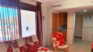 A living room with a sofa, a red coffee table, and a floral armchair. There is a kitchen area with wooden cabinets and a clock on the wall.
