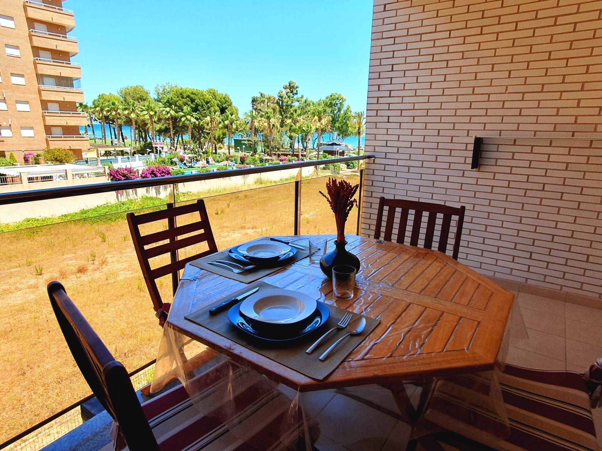 A dining table with chairs on a balcony overlooking a pool area with palm trees and a building in the background.