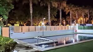 A rectangular swimming pool with a diving board, surrounded by a low wall and palm trees.