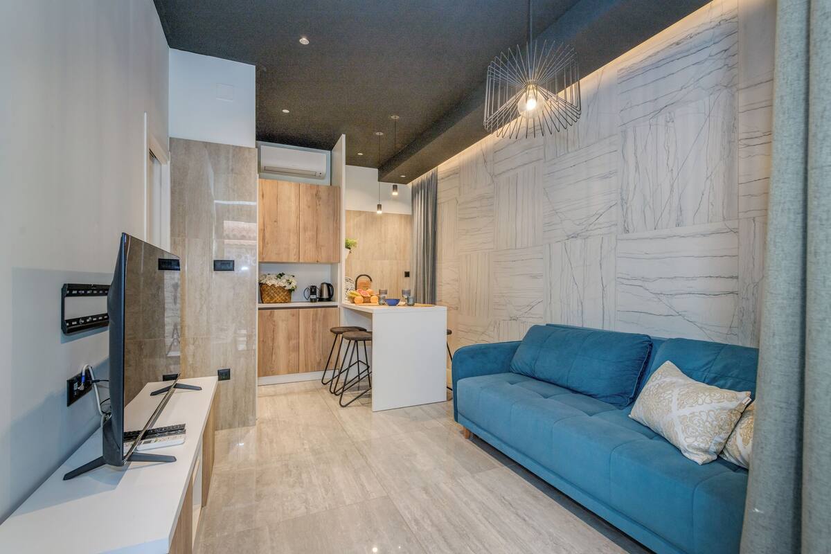 A modern living room with a blue sofa, a flat-screen TV, a kitchen area with wooden cabinets, and a marble wall.
