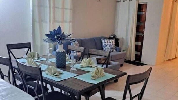 Een eethoek met een rechthoekige tafel voor vier personen, omringd door zwarte stoelen. Op de achtergrond staat een grijze bank en een gestreept kussen.