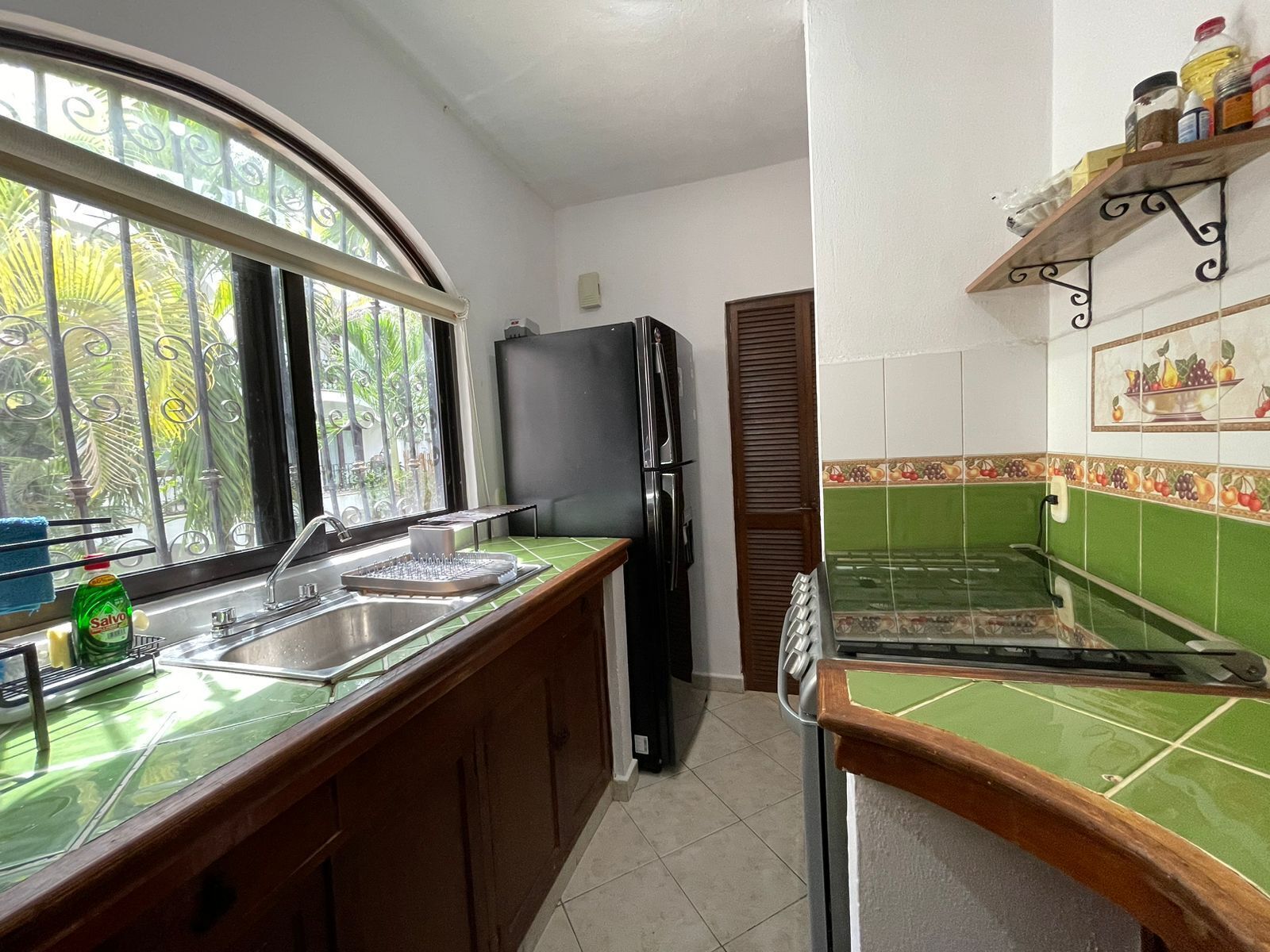 A kitchen with a green countertop, a sink, a refrigerator, and a window with a view of greenery.