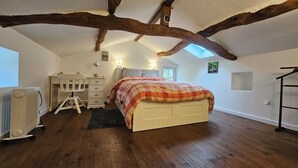 A bedroom with a sloped ceiling, exposed wooden beams, a bed with a plaid blanket, a desk with a chair, a radiator, and a small window.