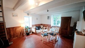 A cozy room with a wooden floor, a dining table with chairs, a sofa, a lamp, and a window with a view of greenery.