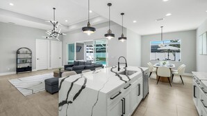 A modern kitchen with a marble island, a dining area with a pool view, and a living room with a sofa and a bookshelf.