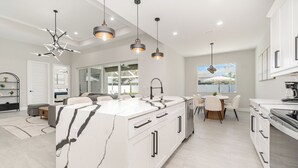 Une cuisine moderne dotée d’un grand îlot central, de luminaires suspendus et offrant une vue sur l’extérieur.