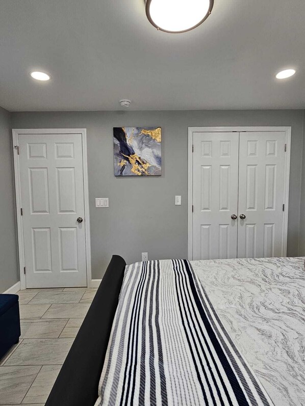 Quarto com uma cama, duas portas brancas, uma luminária no teto e uma obra de arte abstrata na parede.