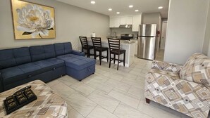 Ein modernes Wohnzimmer mit einem Ecksofa, einem gemusterten Sessel und einem Couchtisch. Im Hintergrund ist ein Essbereich mit einer Küche zu sehen.