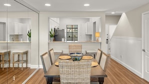 Cocina moderna con armarios blancos, comedor con mesa de madera y sillas de mimbre, y un separador de vidrio.