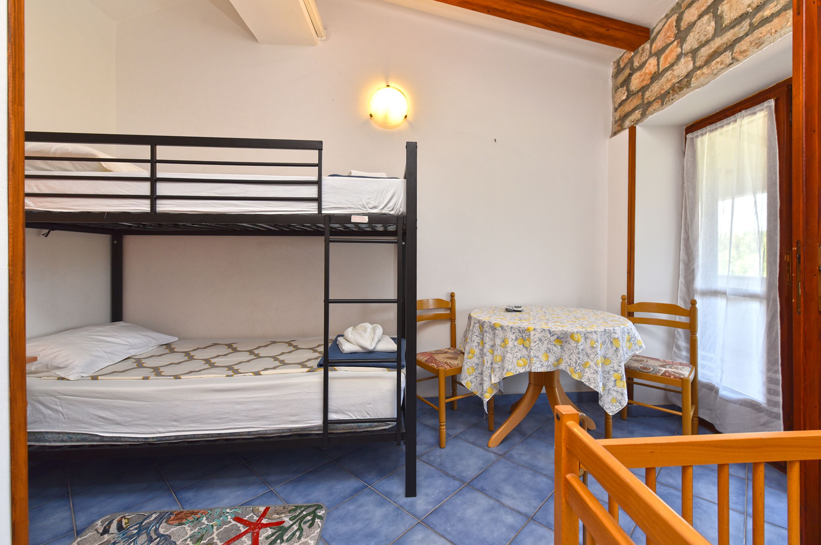 A bunk bed room with a table and chairs, a window with curtains, and a wall-mounted light.