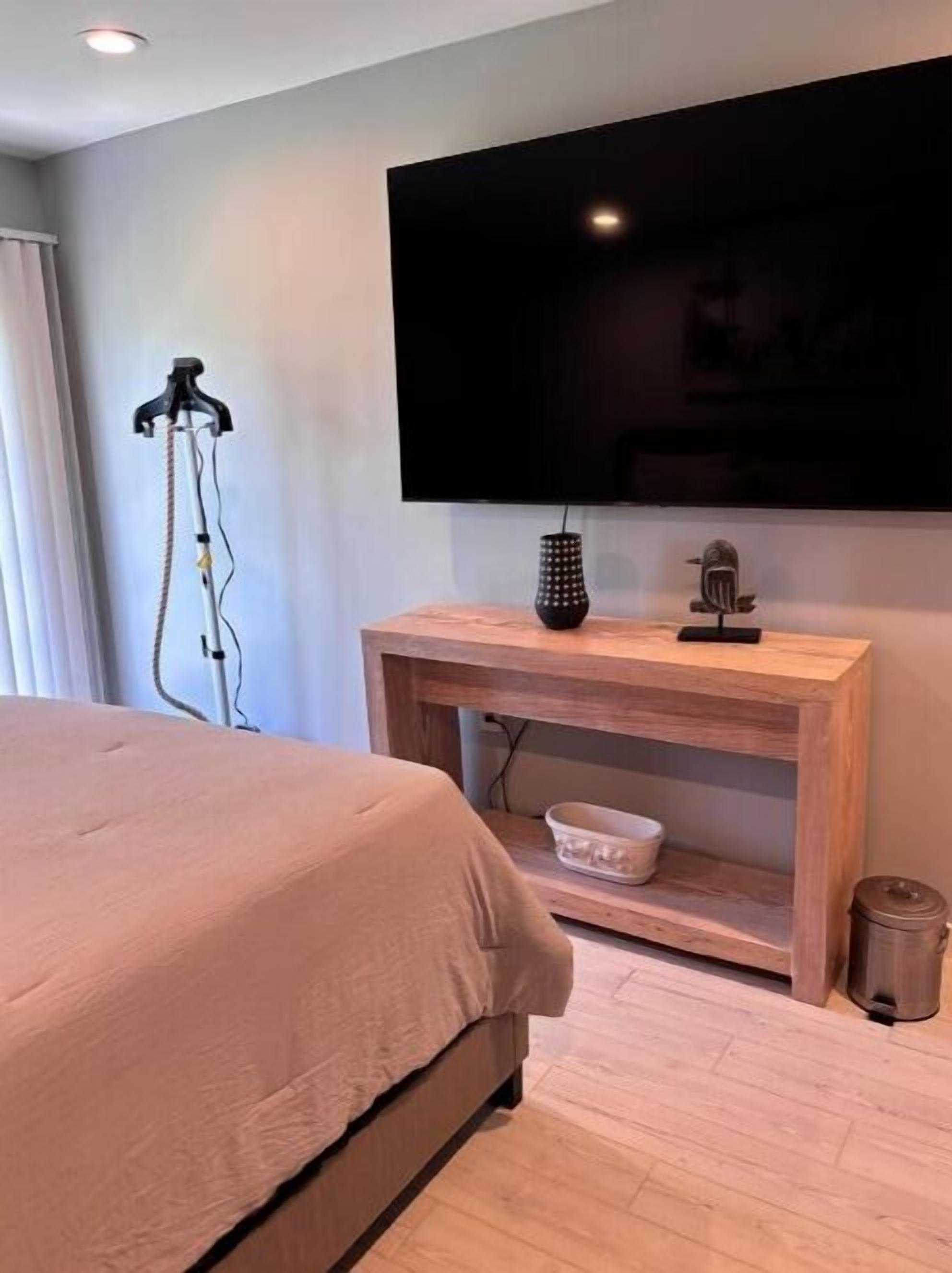 Un dormitorio con cama, televisor montado en la pared, mesita de noche de madera y una mesita auxiliar.