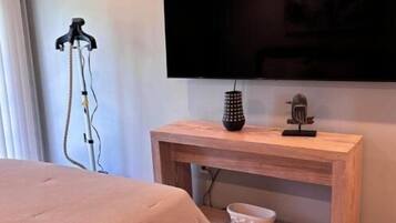 Un dormitorio con cama, televisor montado en la pared, mesita de noche de madera y una mesita auxiliar.