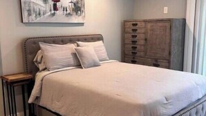 A bedroom with a large bed, a nightstand, a wooden dresser, and a painting on the wall. - Luxury private apartment (Metairie)