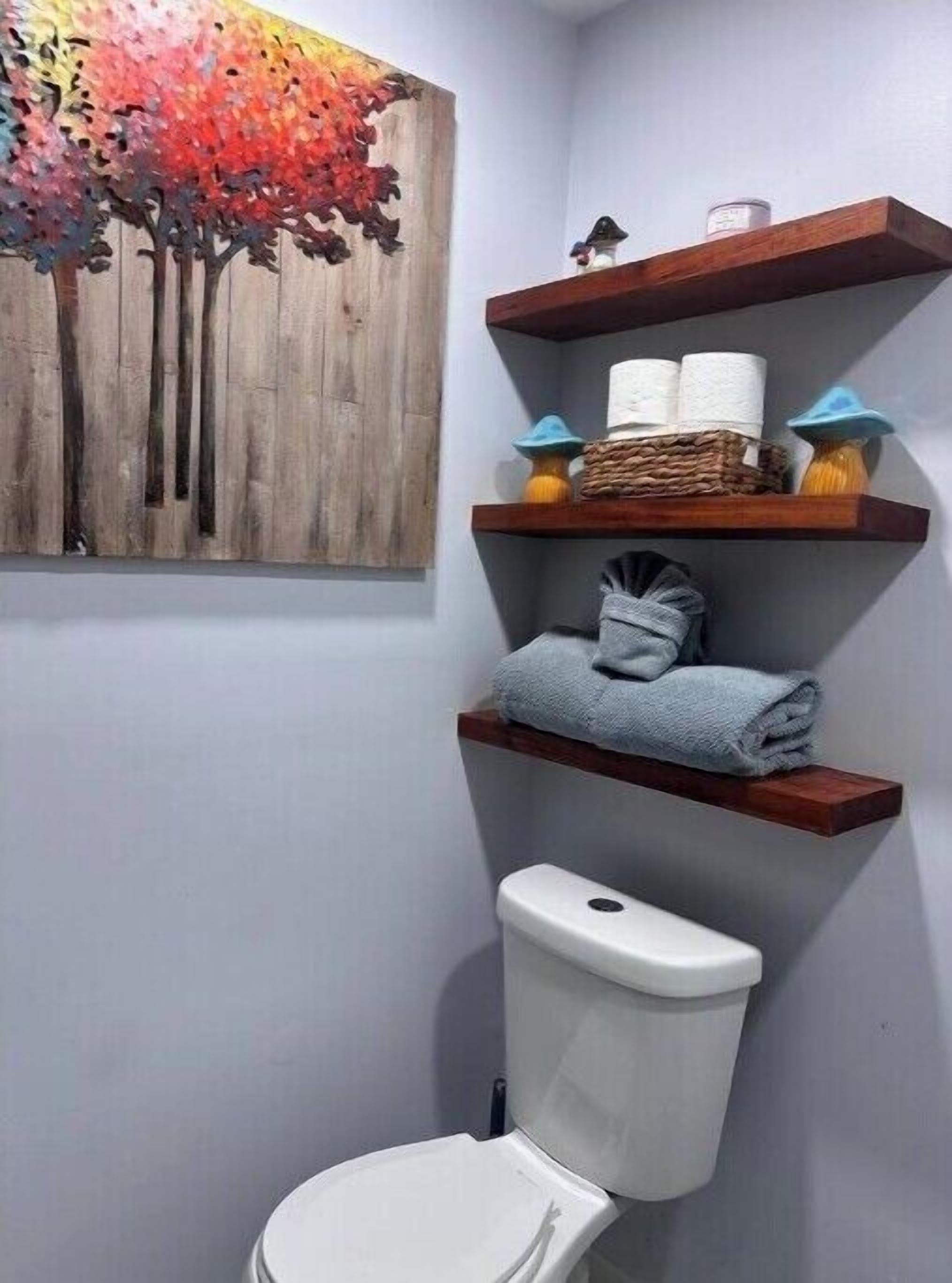 A bathroom with a white toilet, wooden shelves with towels and toiletries, and a decorative wall piece.