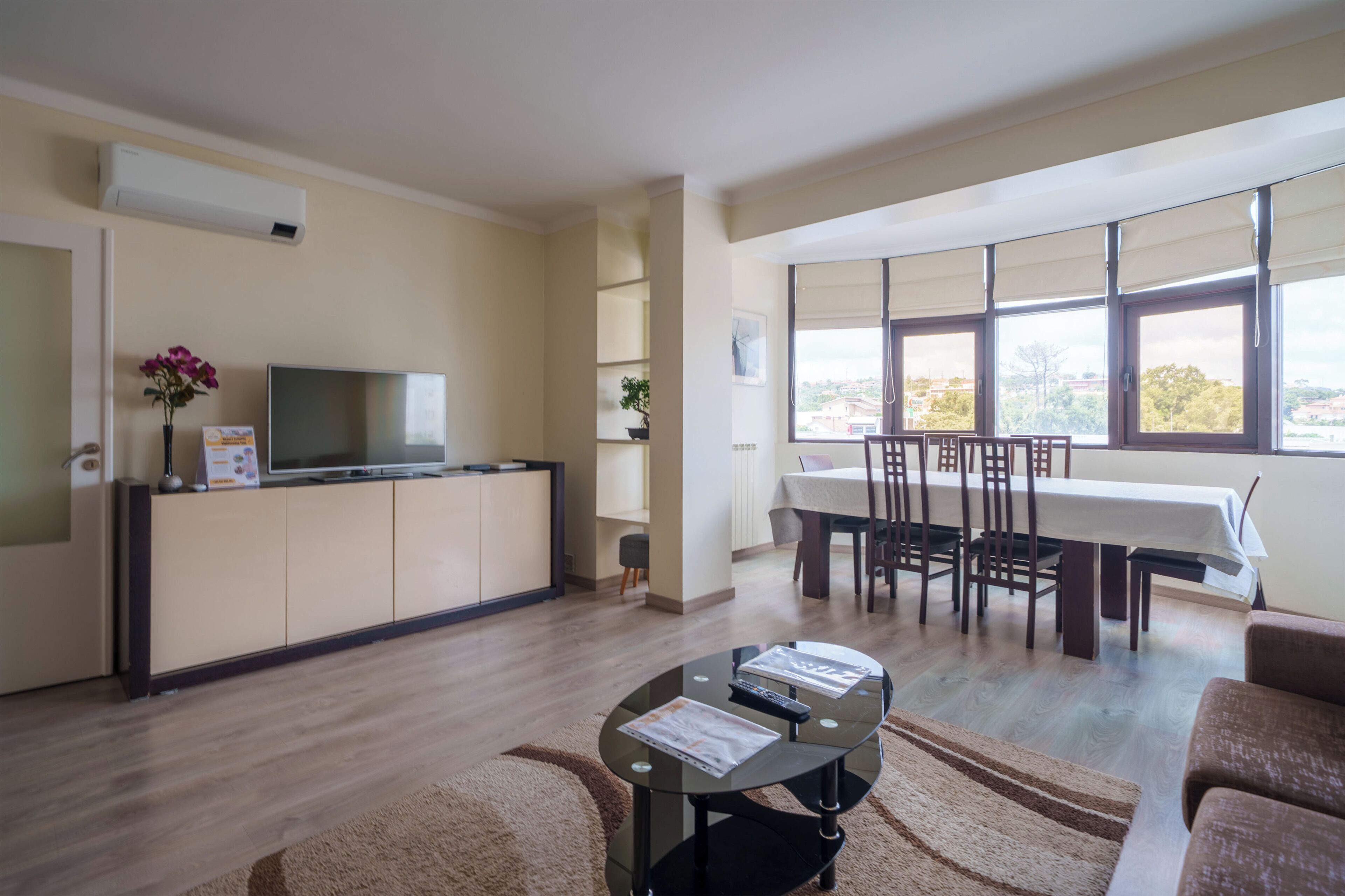 A modern living room with a flat-screen TV, a dining area with a table and chairs, and a large window offering a view.