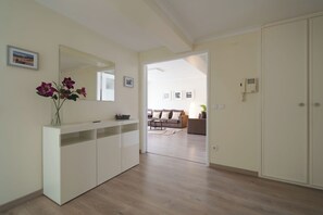 Een modern interieur met een witte console tafel, een spiegel en een vaas met bloemen. Een deur leidt naar een zithoek met een bank en een salontafel.