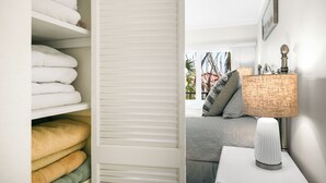 A neatly organized closet with folded towels and a view of a bedroom with a bed, pillows, a lamp, and a window with a view of palm trees. - Cozy & Peaceful Lido Beach Condo — Steps to the Beach and St. Armands.  (Sarasota)