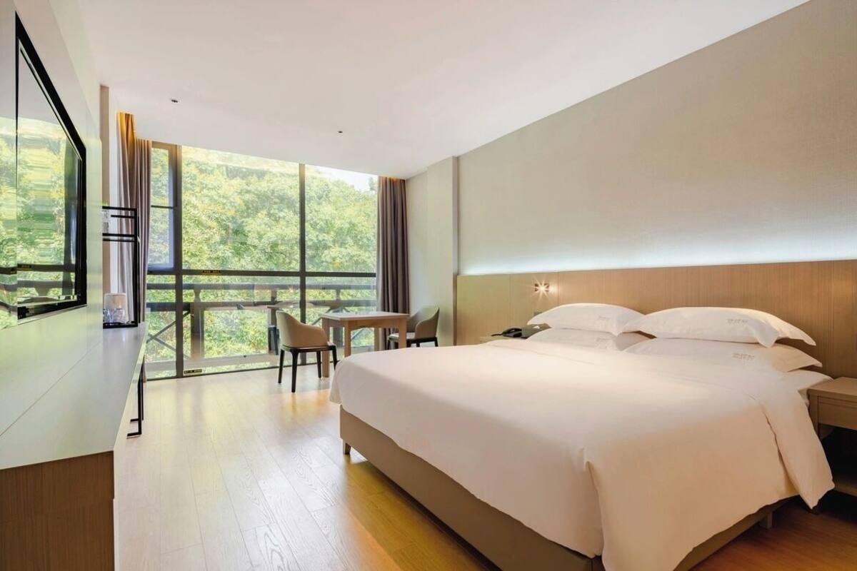 A modern hotel room with a large bed, a desk, and a view of greenery through the windows.
